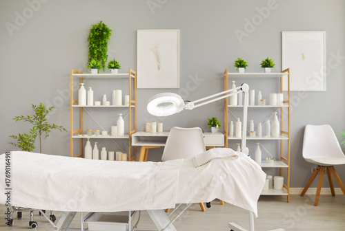 Fotografía Clean and bright spa treatment room with an empty massage bed, towel, magnifying lamp, shelves of cosmetic products