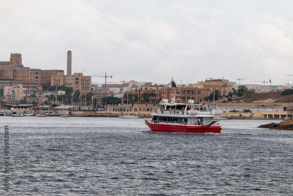 Fototapeta premium A red and white boat sails on the water in front of a city with historical buildings and a tall tower under an overcast sky.