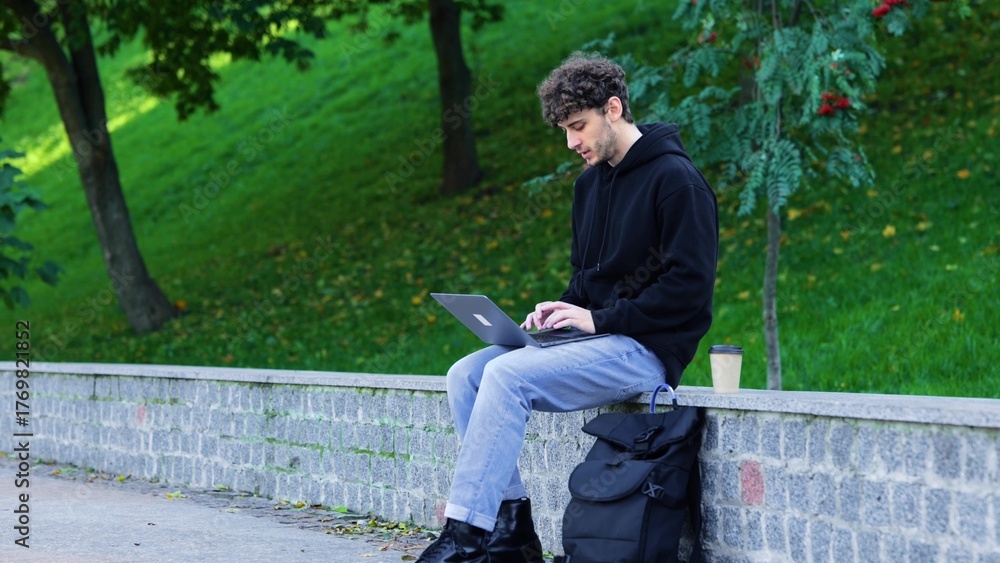 Fototapeta premium Young man sitting in the park and typing on the laptop, coffee next to him. Autumn season. Education, working concept. Slow motion