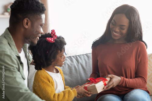 A young girl presents a nicely wrapped gift to her smiling mother while her father looks on. The scene is warm and joyful, capturing a special family moment indoors.