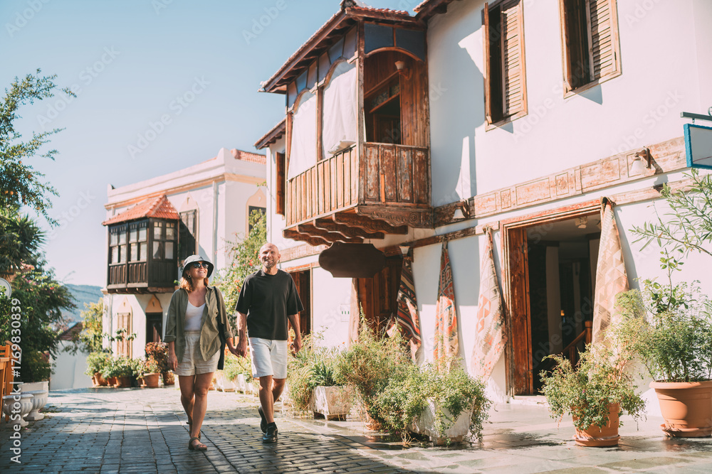 Fototapeta premium Middle aged couple walking hand in hand along narrow street in Kas town historical center. Romantic travel, love relationship, honeymoon vacation and happy retirement lifestyle concept.