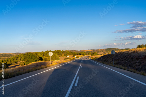 Empty road winds through dry, sunlit hills under blue sky.