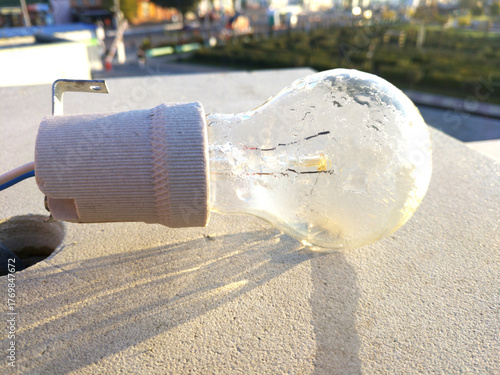 The light bulb hangs against a light concrete wall, illuminated by sunlight. Subtle shadows and traces of time are visible on the surface, creating an atmosphere of simplicity and everyday life.