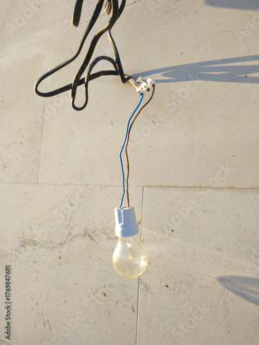 The light bulb hangs against a light concrete wall, illuminated by sunlight. Subtle shadows and traces of time are visible on the surface, creating an atmosphere of simplicity and everyday life.