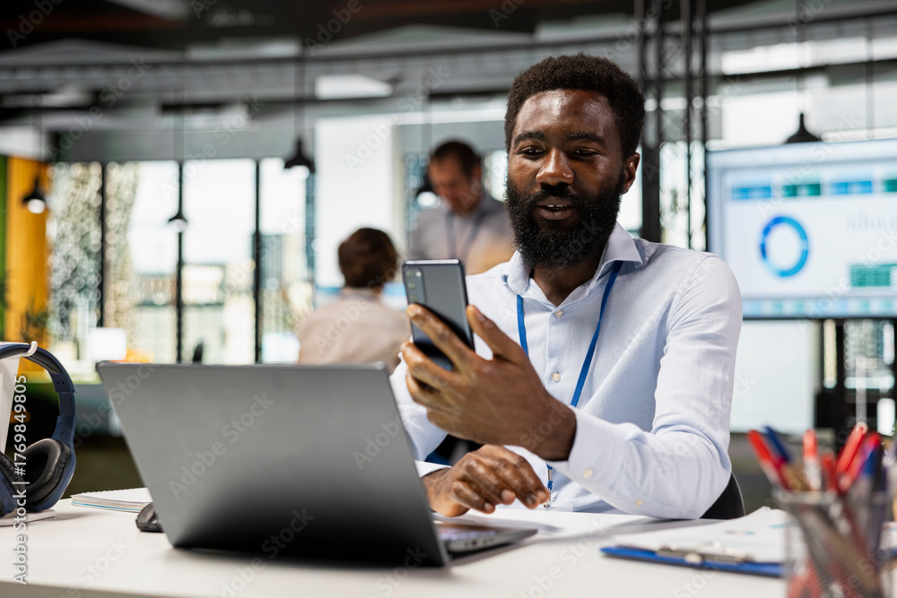 Fototapeta premium African american project manager sitting with cellphone in hand, discussing with client on video call. Black male employee in modern office, taking short break to speak with family on mobile device.