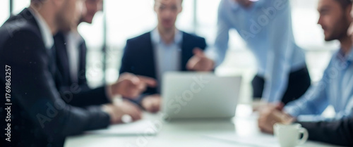 Business meeting with blurred figures around a table and laptop, illustrating teamwork, strategy, and creative discussion among professionals in a bright corporate environment.