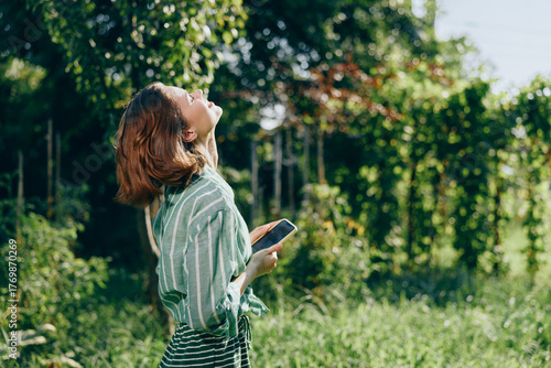 A woman stands in a sunny meadow, looking upward with a phone in hand, surrounded by lush greenery and warm light.