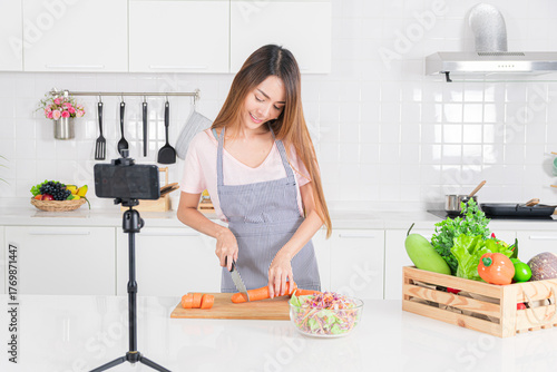 Young asian woman chopping carrot while filming a cooking demonstration in her bright white kitchen emphasizing healthy eating.