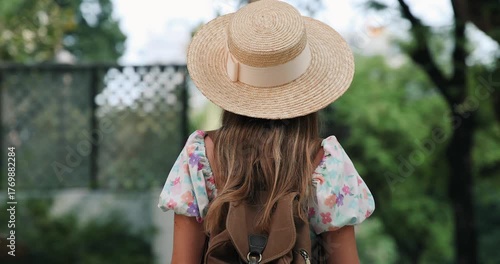 Woman wearing straw hat with floral dress, backpack in park. Outdoor leisure, summer fashion, nature exploration.