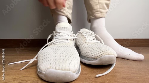 Close up of a woman putting on white sneakers and tying shoelaces indoors. Preparing for fitness, workout, jogging, training, or casual activity on wooden floor