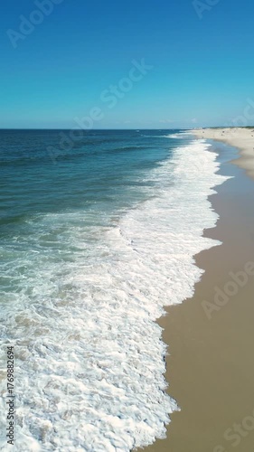 Aerial drone video of the Atlantic Ocean shoreline with gentle waves rolling onto a sandy beach under a clear blue sky. Peaceful summer seascape perfect for travel and nature-themed projects.