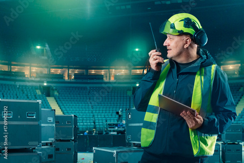 Obraz na plátne Stage supervisor in safety gear communicates via walkie-talkie while holding tablet during concert equipment setup in arena