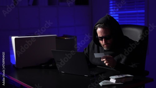 Hacker using laptop in dark room. A man wearing a hoodie and sunglasses sits in a dark room with a laptop, holding a credit card under low light. The scene cybercrime, theft, or online fraud.