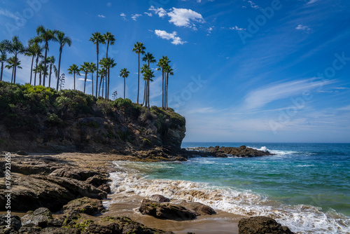 Rocky cliffs, palm trees, turquoise water, blue sky and a sandy beach. Crescent Bay in Southern California is one of the most beautiful beaches in Orange County.