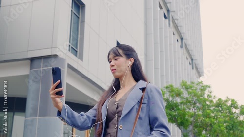 Young business businesswoman happy outdoor walking city street, enjoying music on smartphone, traveling near modern office building during bright day.