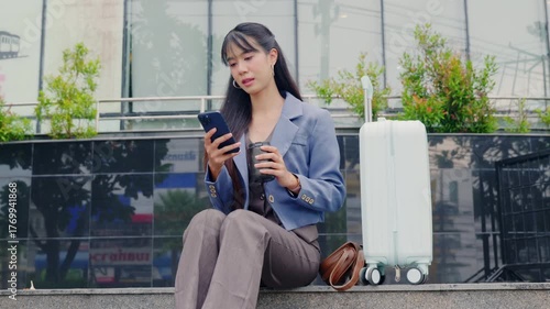 Young business businesswoman enjoys outdoor travel, connecting using mobile phone, sipping coffee, relaxing during busy urban trip, sitting composedly near modern office building with luggage ready.