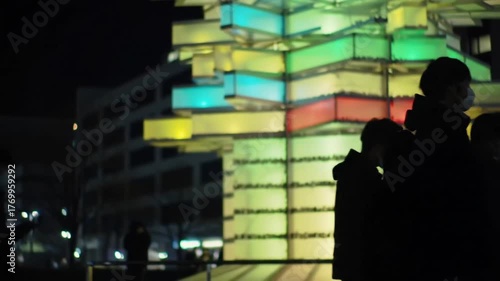People wearing masks walking past a colorful building at night in an urban area
