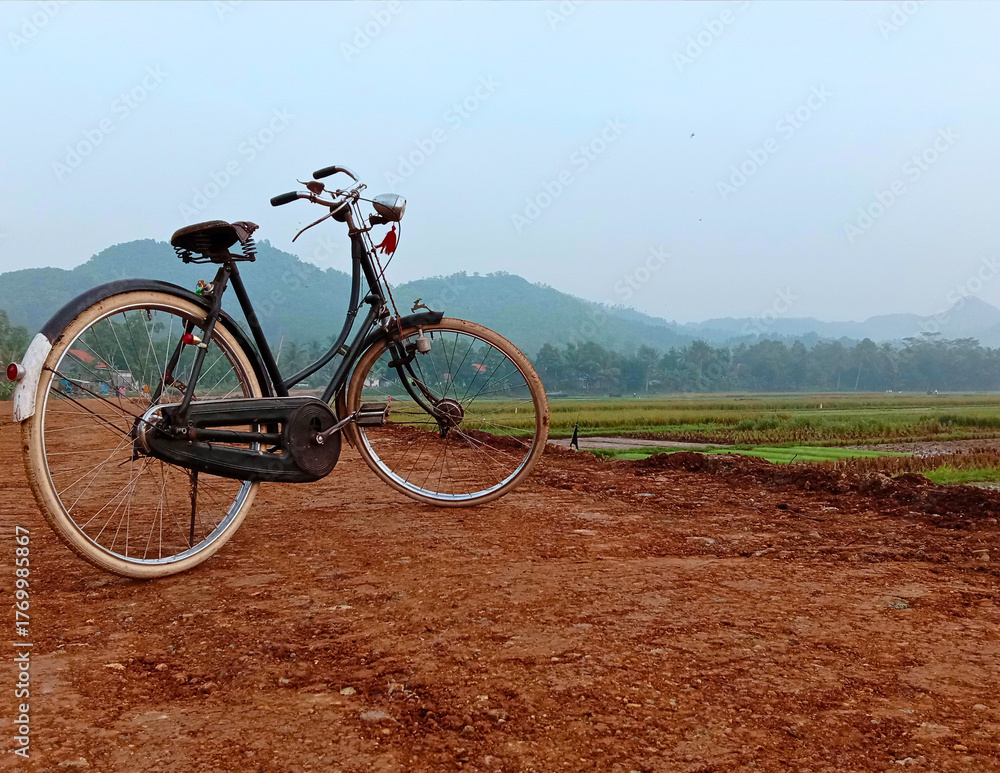 Fototapeta premium Vintage bicycle in rice field. Classic old road bike on rural dirt road with mountain background. Sustainable travel, country lifestyle, and World Bicycle Day 2026 concept in tropical landscape.