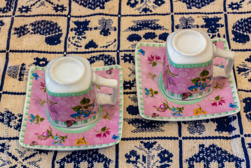 Floral porcelain cup upside down on a square saucer, resting on an embroidered tablecloth with geometric and traditional patterns, symbolizing Brazilian craft and domestic culture.
