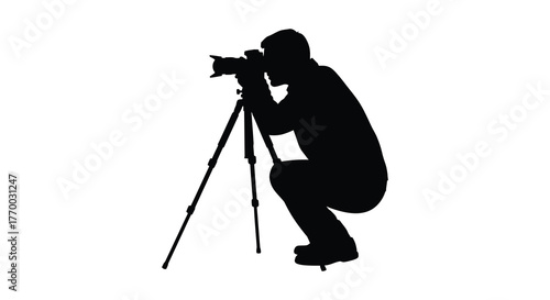 A photographer crouches low to the ground using a professional camera mounted on a tripod captured in a striking black silhouette against a white background silhouette