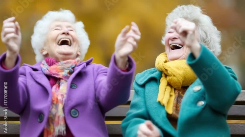 Joyful healthy centenarian elderly woman senior friends laughing on bench celebrating autumn outing in colorful coats and scarves