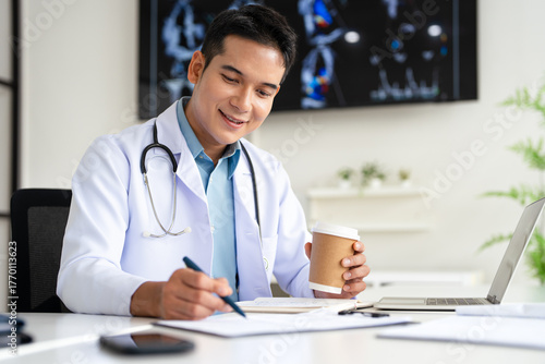 Professional doctor holding a hot coffee drink cup.doing paperwork checking medical documents at workplace. Concentrated physician reading medic form analyzing patient diagnosis or report in hospital.