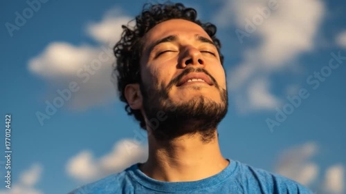 A young man with curly hair meditates outdoors under a cloudy blue sky