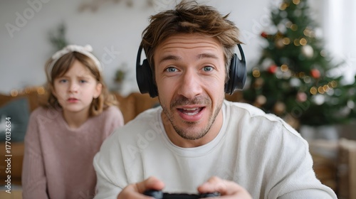 A man wearing headphones excitedly plays a video game while his daughter looks on with a grumpy expression near a Christmas tree