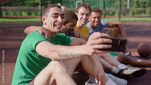 Smiling sporty fit diverse multiracial male basketball players sitting on outdoor court, taking selfie shot on cellphone, expressing cheerful mood and positivity while relaxing after game.
