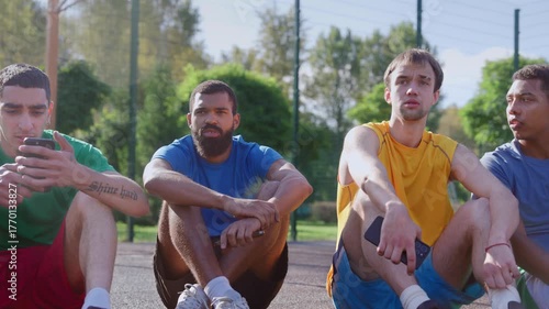 Positive sporty fit diverse multiracial male athletes sitting on outdoor basketball courts friendly talking and sharing while streetball players relaxing after game.