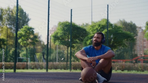 Portrait of sporty fit bearded African American male basketball player in wireless headphones sitting on outdoor court, enjoying music while taking rest after sports training in morning.