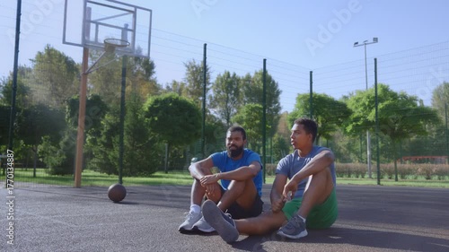 Attractive sporty fit African American basketball players sitting on outdoor court, communicating and discussing match while friends relaxing after streetball game .