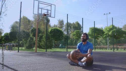Positive athletic bearded African American male basketball player sitting on outdoor court, wearing wireless headphones and enjoying music while relaxing after streetball training.