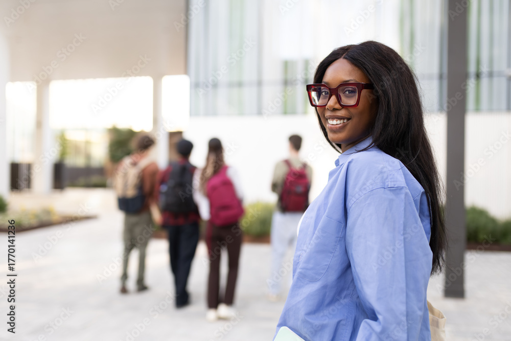 Fototapeta premium Smiling university student turning around on campus with friends walking in background