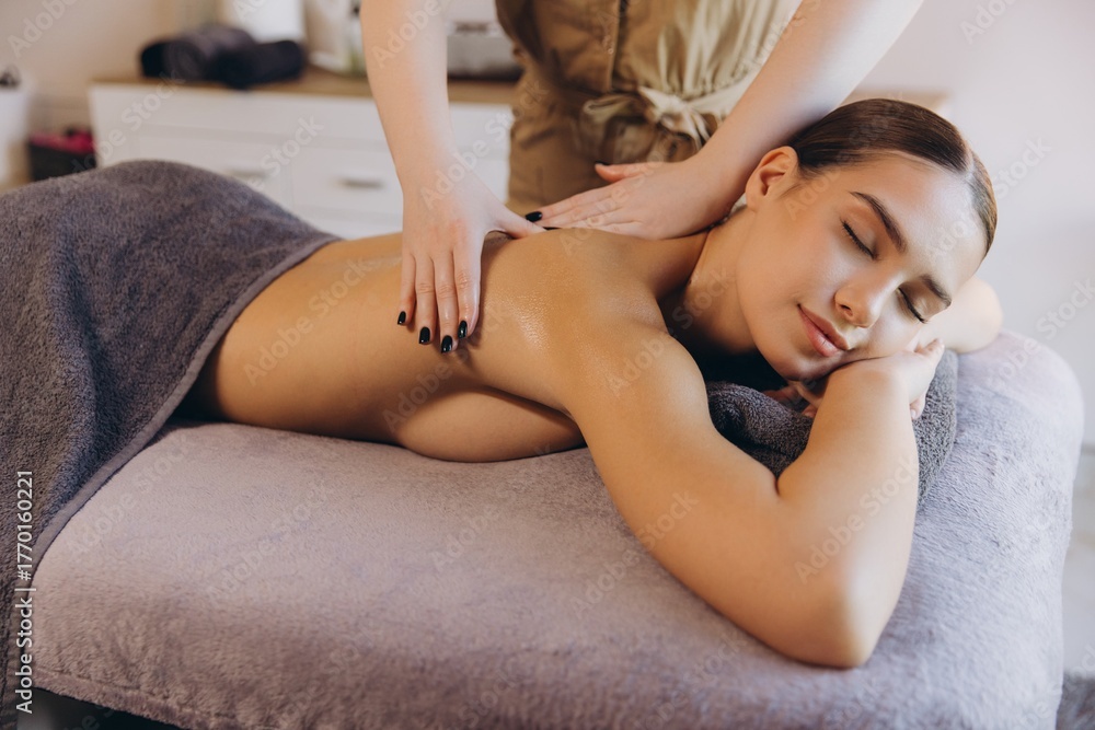 Obraz premium Professional masseuse applying massage oil on back of young woman lying on massage table in spa salon