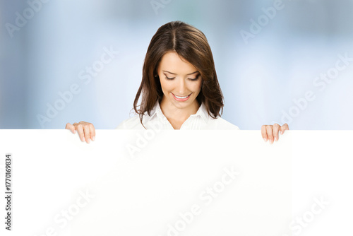 Happy smile woman wear corporate shirt peep out show pointing advertise blank white banner signboard with empty sign text free space, stand against blurred office background Business ad concept
