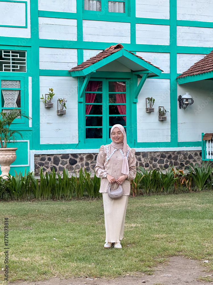 Φωτογραφία A woman in a light outfit and headscarf poses by a colorful house and lush green