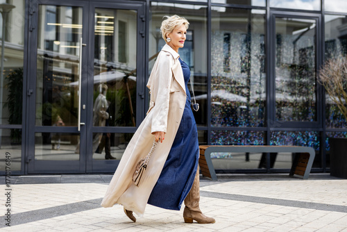 Beautiful Woman Wearing Fashionable Spring Or Fall Clothes, beige trench coat, denim dress and high boots. Female stylish Model walking Street. Autumn trend street fashion style. Outdoors