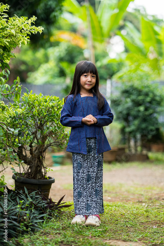 A young girl wearing traditional kebaya and batik skirt standing outdoors, representing Indonesian traditional clothing and culture.