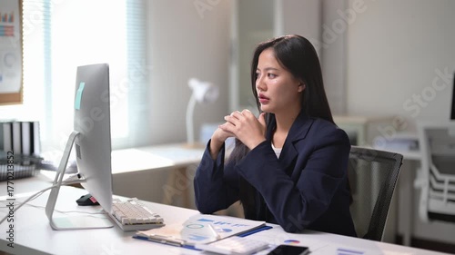 A woman is sitting at a desk in front of a computer monitor. She is wearing a black suit and she is typing. The room is well-lit and has a professional atmosphere