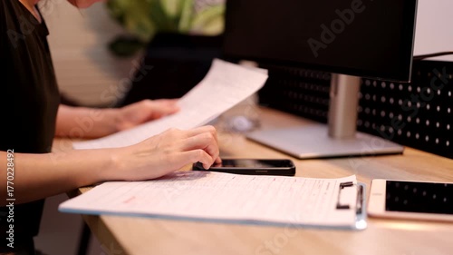 A person reviews tax documents and calculates financial data using a smartphone and computer at a desk.