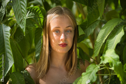 Portrait of a young woman, blonde, blue eyes, slim, wearing a green dress, looking sweetly and tenderly at camera, surrounded by green plants. Concept: femininity, innocence, youth.