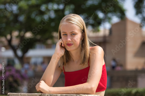 Portrait of a young woman, blonde, blue eyes, slim, wearing a red top, looking thoughtfully at camera, leaning against a stone wall. Concept: femininity, innocence, youth.