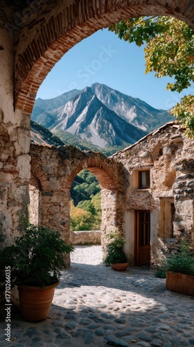 Ancient Stone Arches Frame a Majestic Mountain View in a Historic Village Courtyard with Cobblestone Pavement and Lush Greenery on a Sunny Day