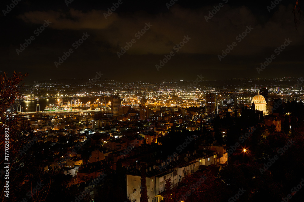 Fototapeta premium Night panoramic view of Haifa with bright port lights, city skyline, and illuminated Baha'i Gardens overlooking the bay.