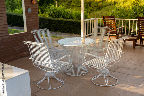 White metal patio furniture set featuring a round table and swivel chairs, surrounded by lush greenery and shaded by a roof. Perfect for concepts of outdoor living, garden design, resort seating