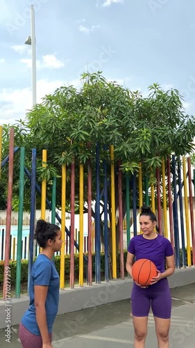 Two young women in sportswear having a conversation while one holds a basketball on an outdoor court