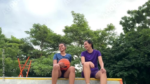 Two young women athletes sitting together and talking at an outdoor basketball court
