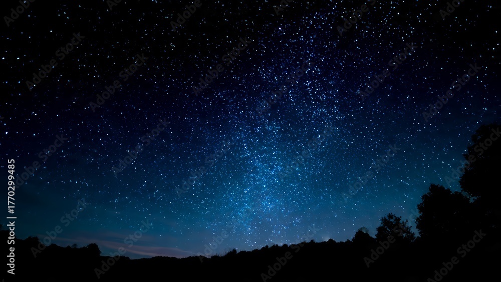 Fototapeta premium A vast expanse of twinkling stars filling the deep blue night sky above dark silhouetted trees and hills