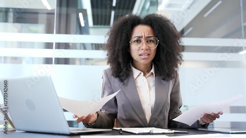 Disappointed african american businesswoman having difficulty with paperwork sitting in business office looking at camera. Puzzled frustrated female looks at documents and cannot understand problem